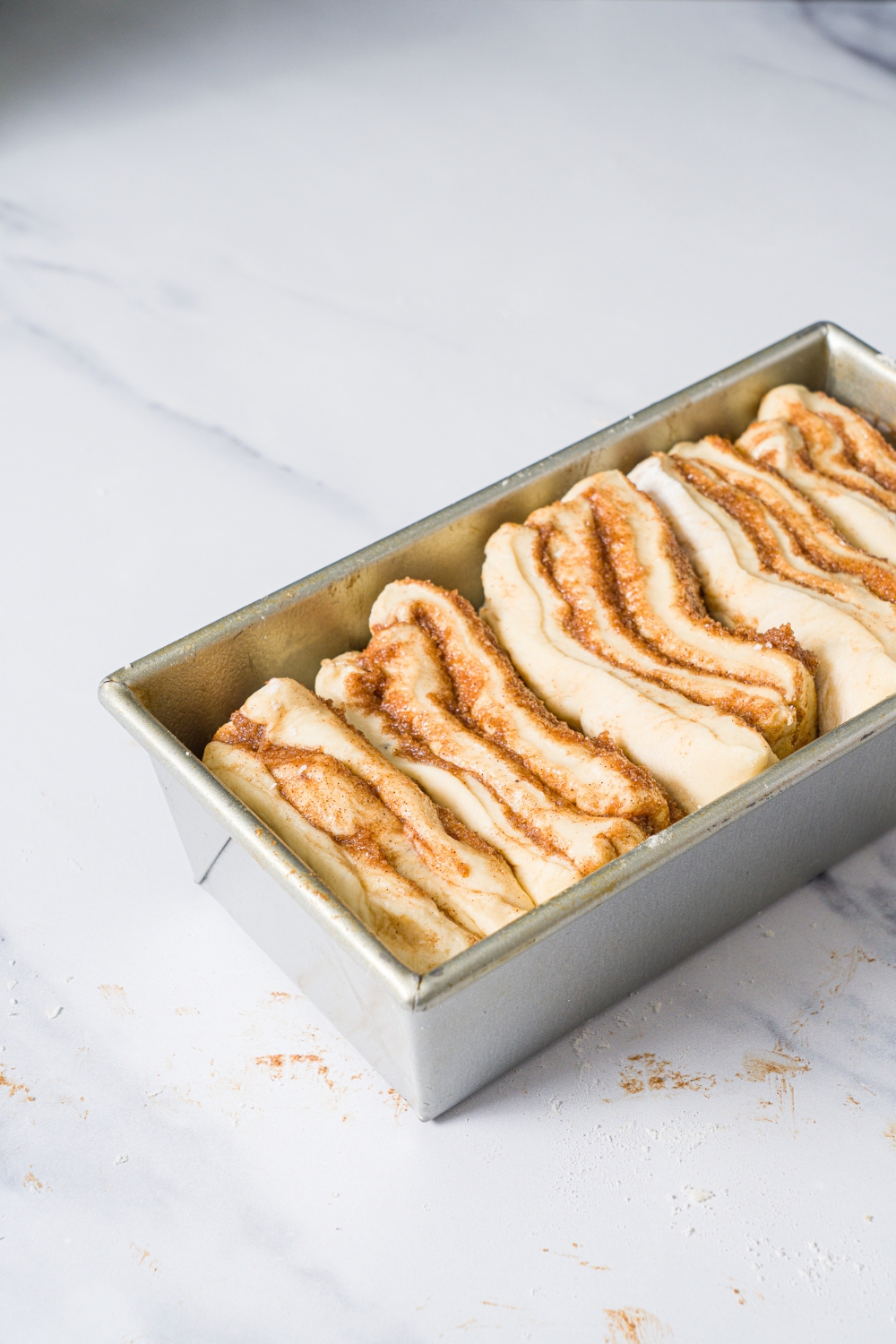 A loaf pan with shaped cinnamon pull apart dough pieces rising in the pan. The pan is on a marble counter.