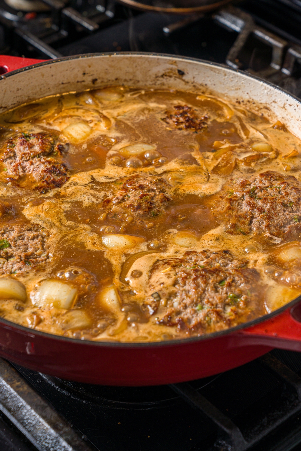A cast iron skillet with hamburger steaks cooking in onion gravy. The skillet is cooking on a stove.