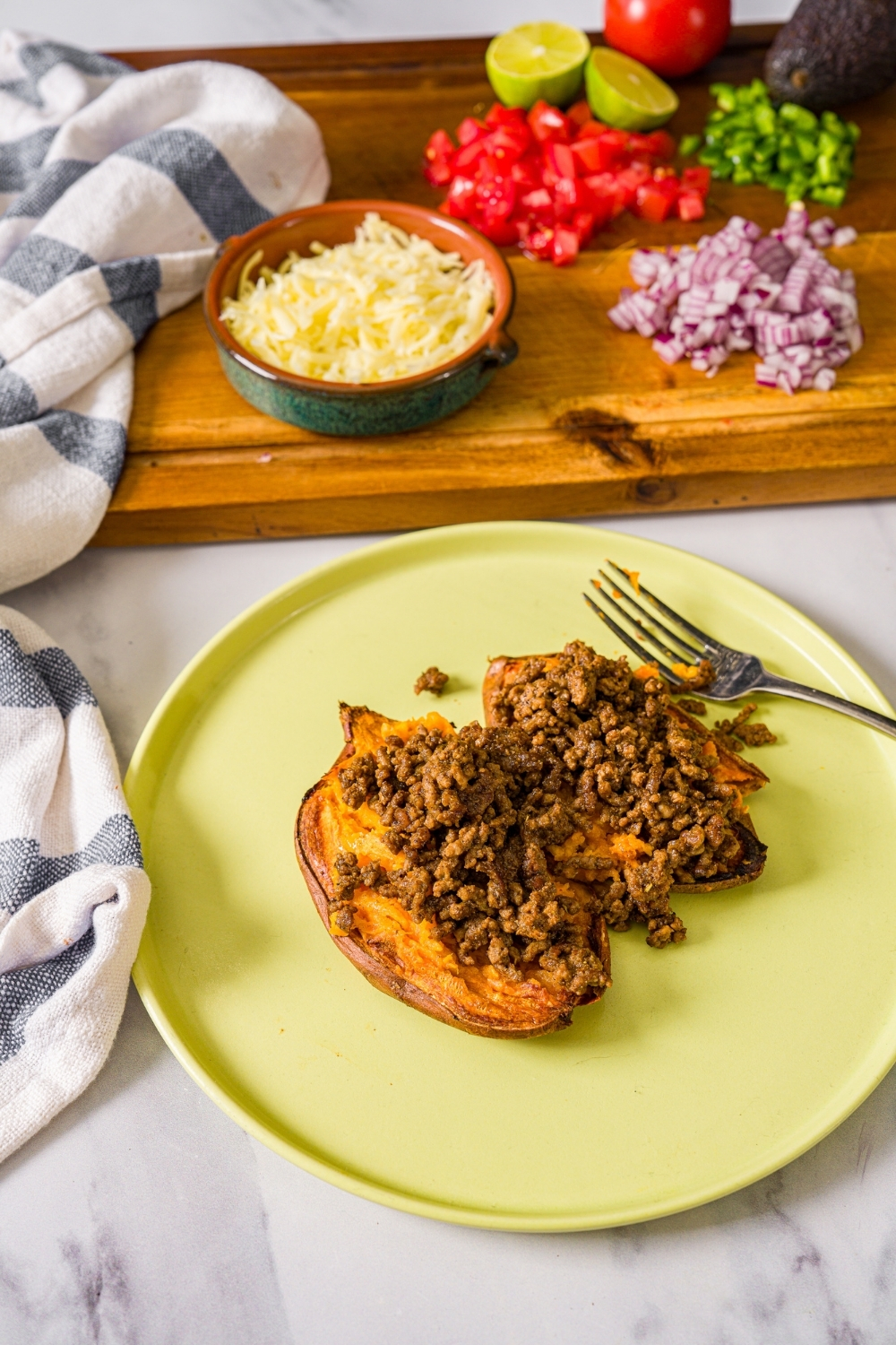 A green plate with an open baked sweet potato topped with seasoned taco meat. The plate is on a marble counter with a board of toppings including shredded cheese, and chopped vegetables.