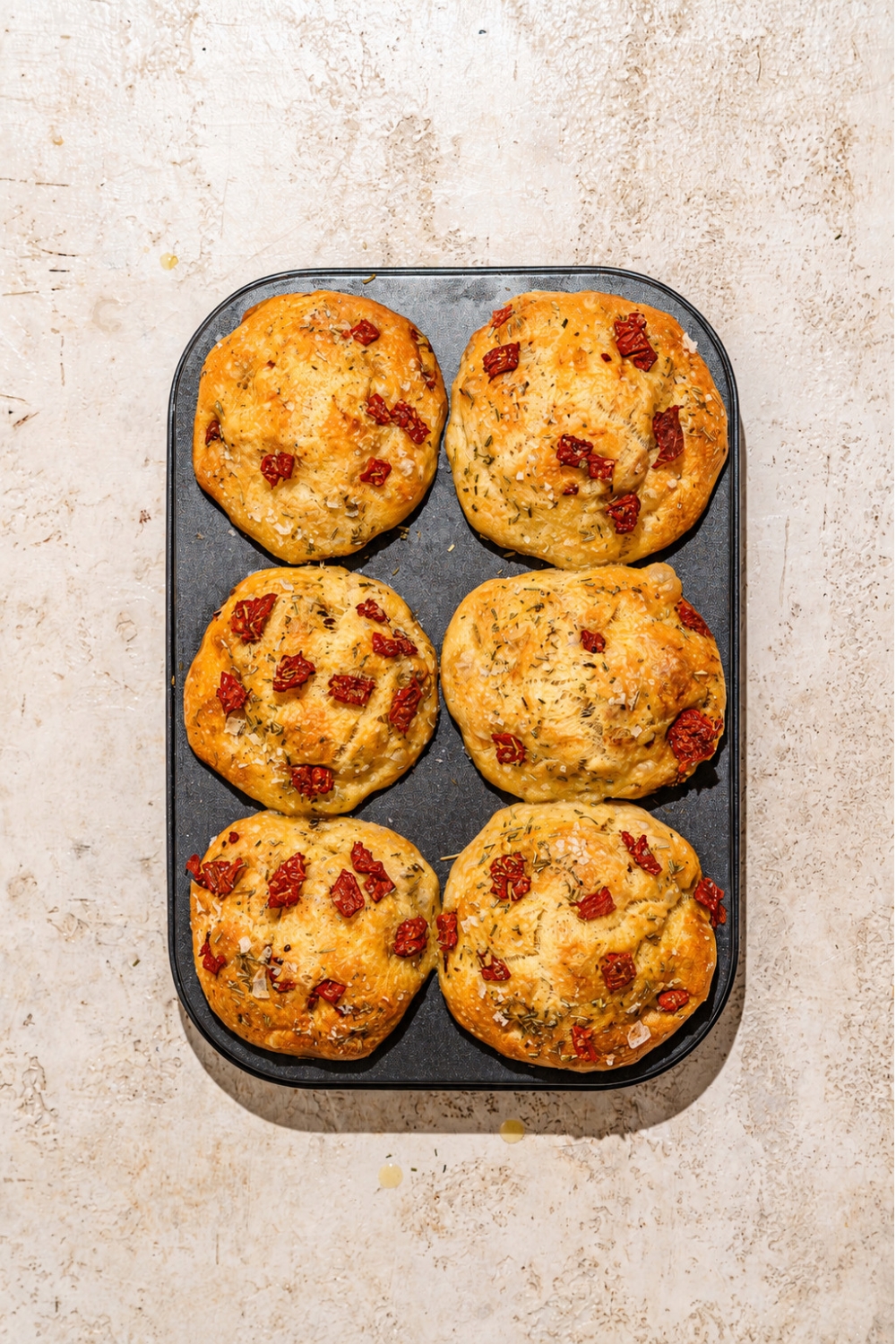 A muffin tin with baked rosemary focaccia muffins. The tin is on a tan counter.