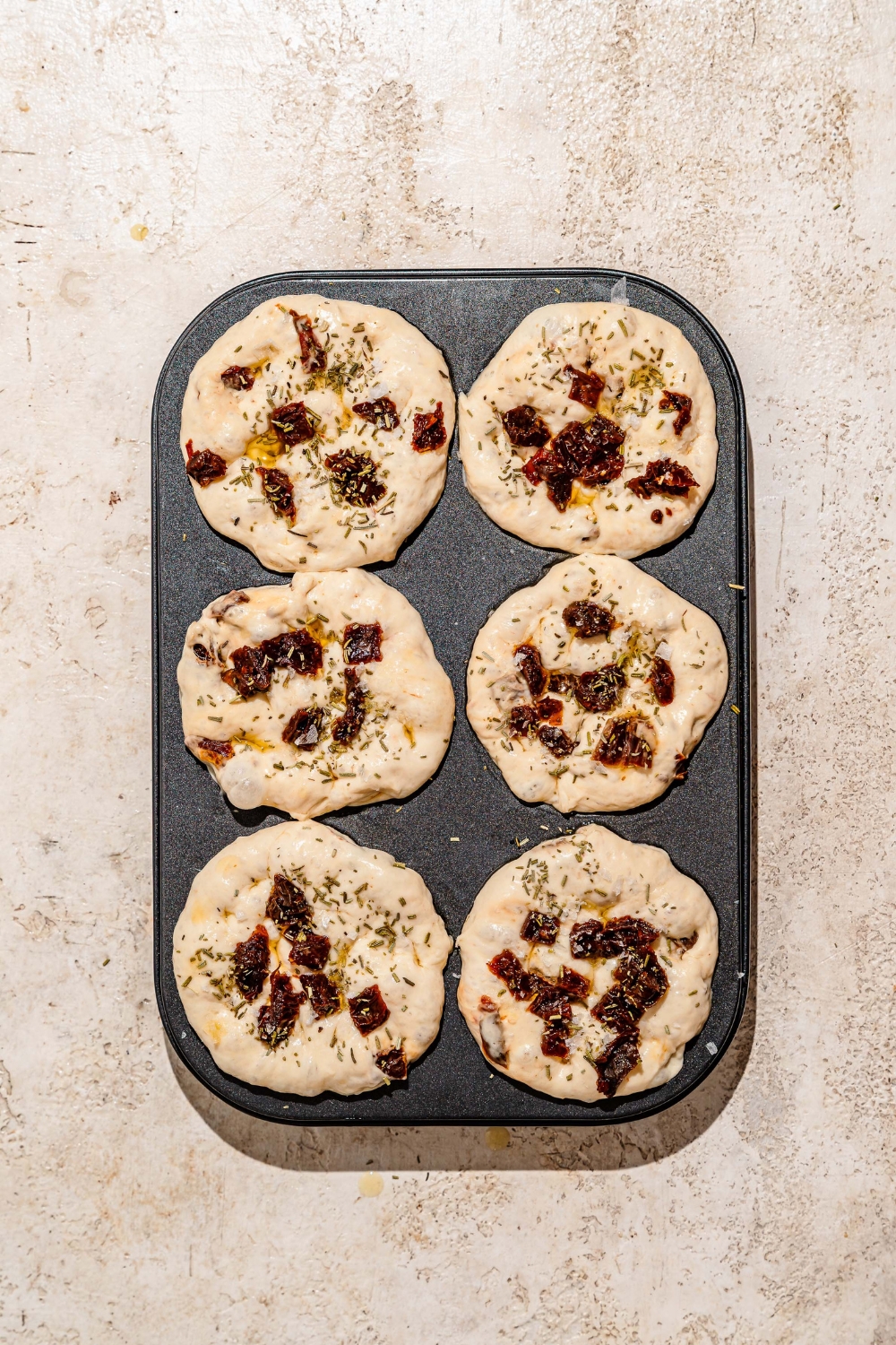 A muffin tin with focaccia dough topped with oil and rosemary. The tin is on a tan counter.