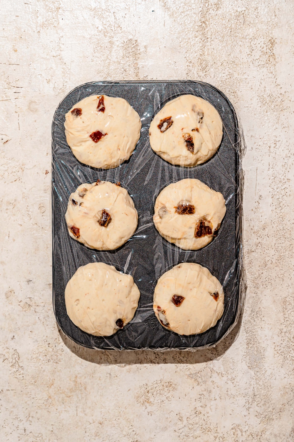 A muffin tin with focaccia dough. The tin is wrapped in saran wrap and on a tan counter.