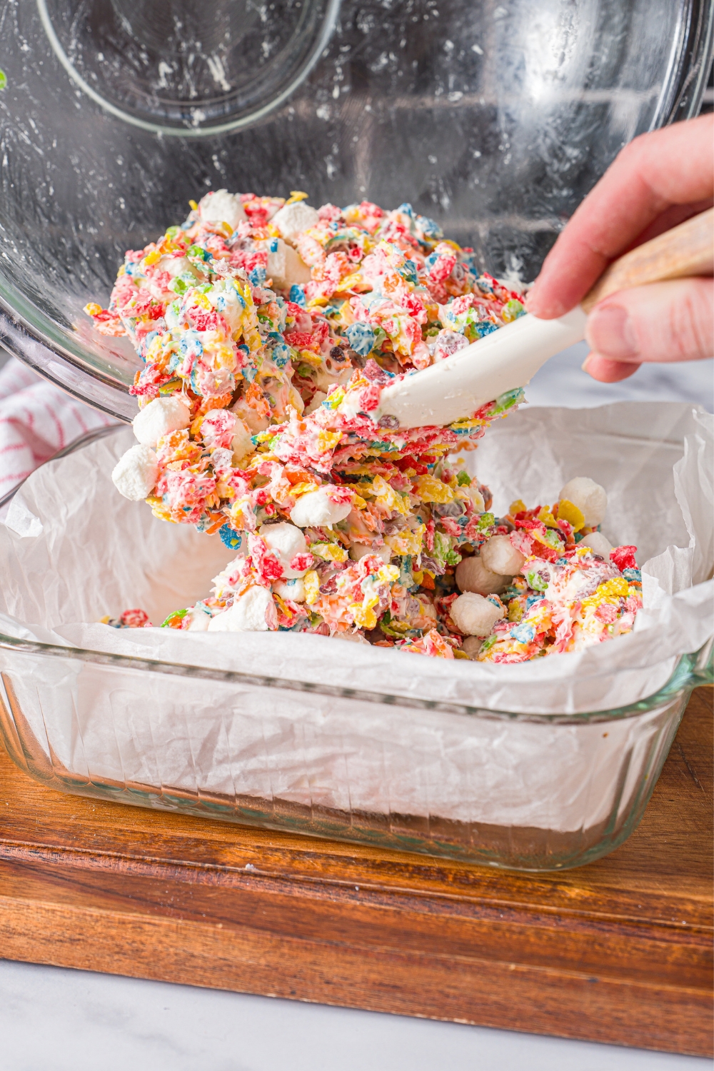 A spatula spreading a fruity pebble rice crispy mixture from a bowl into a baking dish lined with parchment paper.