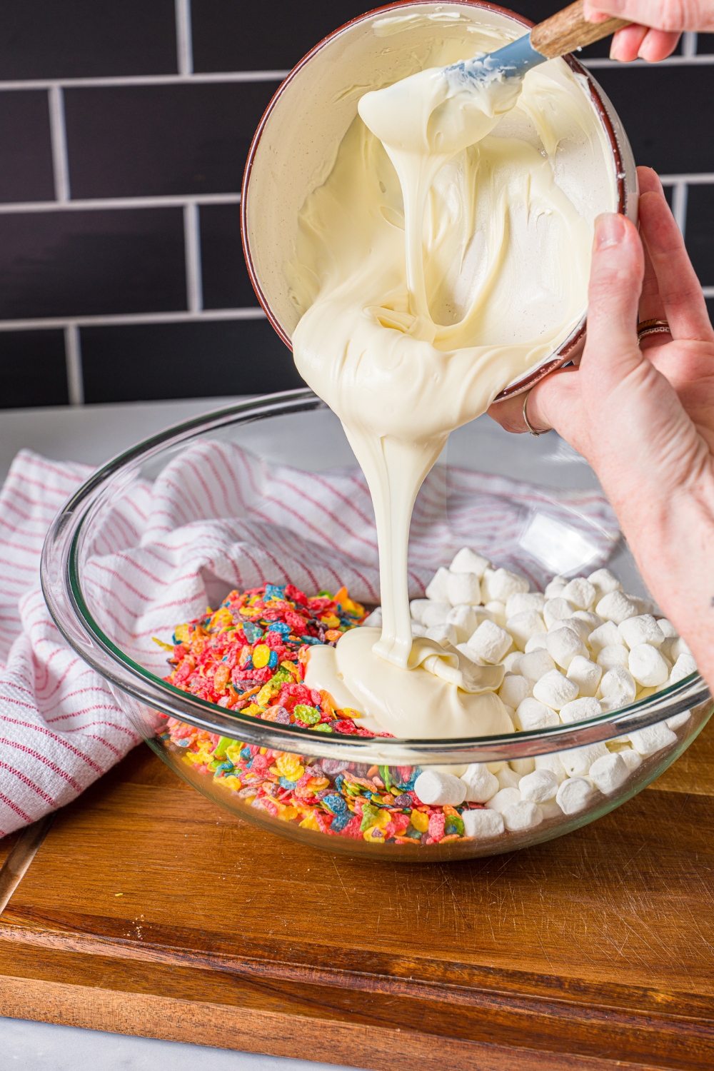 A bowl of melted white chocolate being poured into a bowl of fruity pebbles and marshmallows. The bowl is on a wooden board.