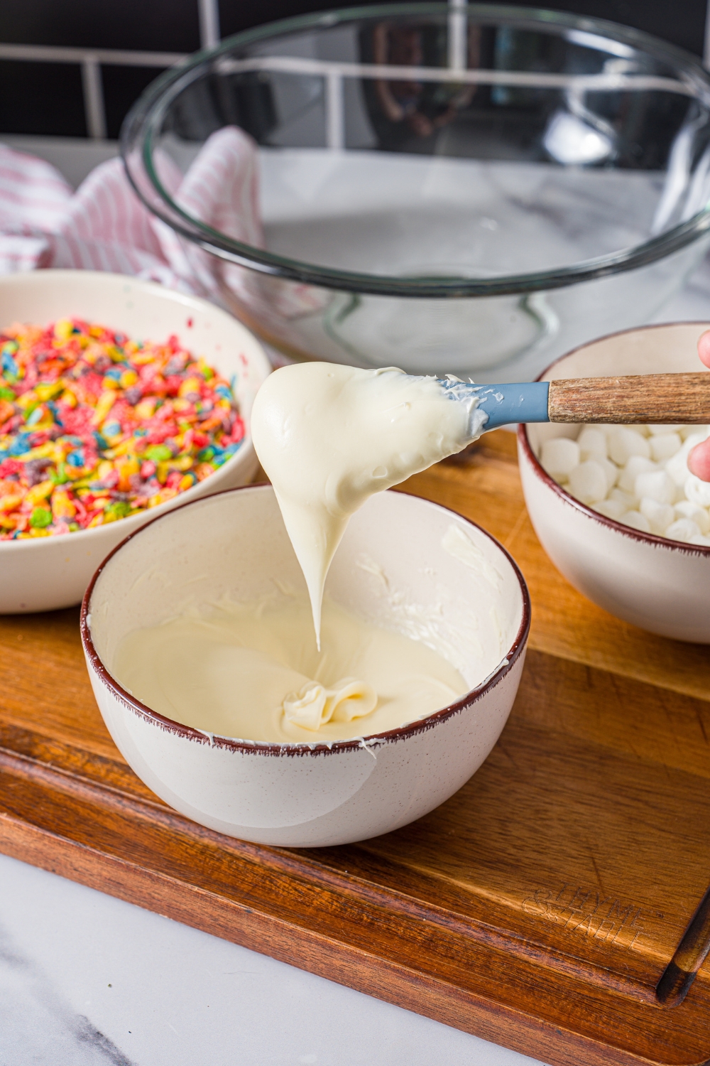 A spatula dipped in a bowl of melted white chocolate. The bowl is on a wooden board with a bowl of fruity pebbles.