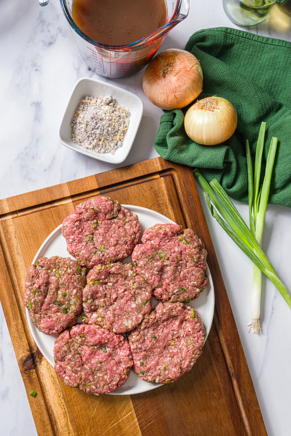 A plate of seasoned hamburger steak patties on a wooden board. The board is on a marble counter with additional ingredients.