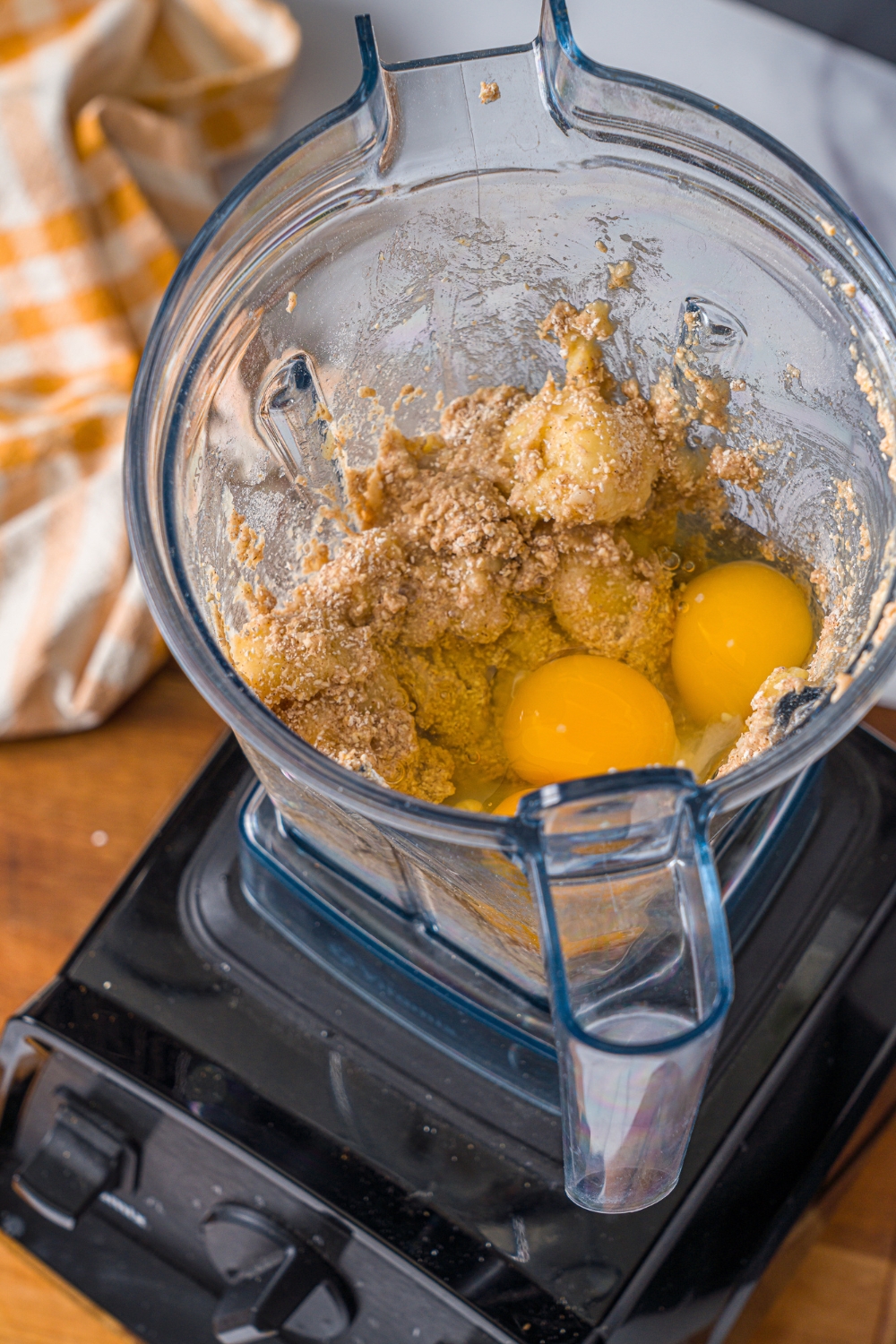 A blender with egg, vanilla, and a dry ingredient mixture for banana protein muffins. The blender is on a wooden board.
