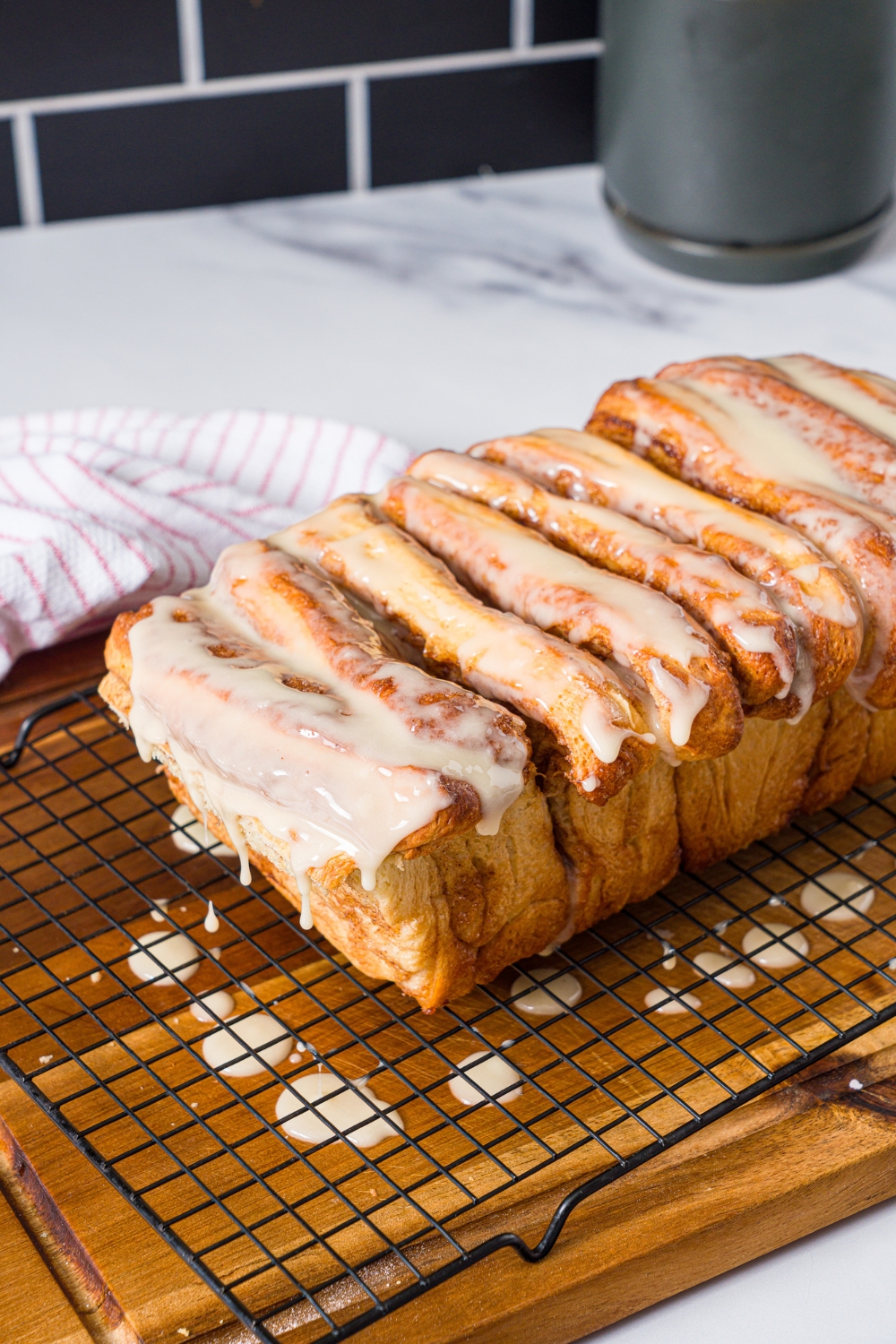 A loaf of cinnamon pull apart bread topped with glaze on a wire rack on a wooden board. The board is on a marble counter with a red striped napkin.