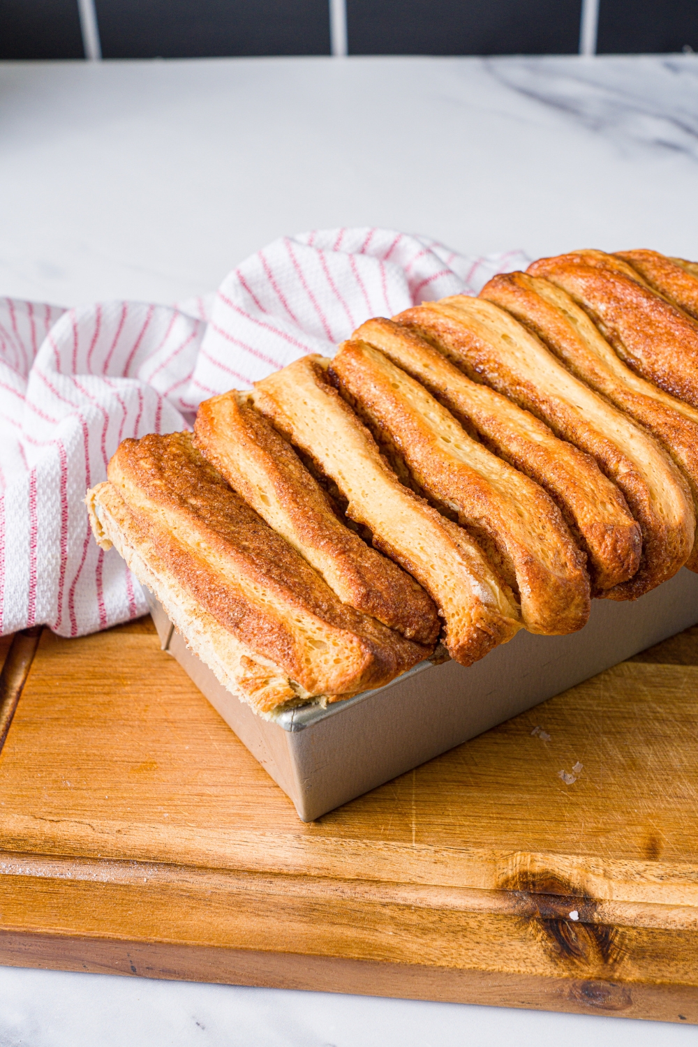 A loaf pan with baked cinnamon pull apart bread. The pan is on a wooden board with a red striped napkin.
