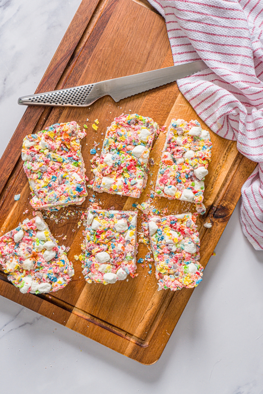 A wooden board with sliced fruity pebbles rice crispy treats with a knife and red striped napkin.
