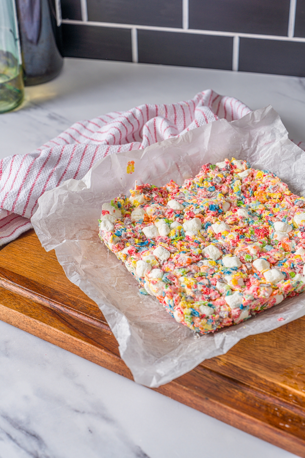 A wooden board with a brick of fruity pebbles rice crispy treats on a piece of parchment paper. The board is on a marble counter with a red striped napkin.