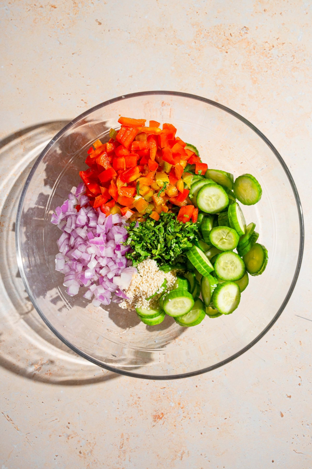 A large glass bowl with sliced cucumbers, diced red bell pepper, chopped red onion, cilantro, and sesame seeds. The bowl is on a tan counter.