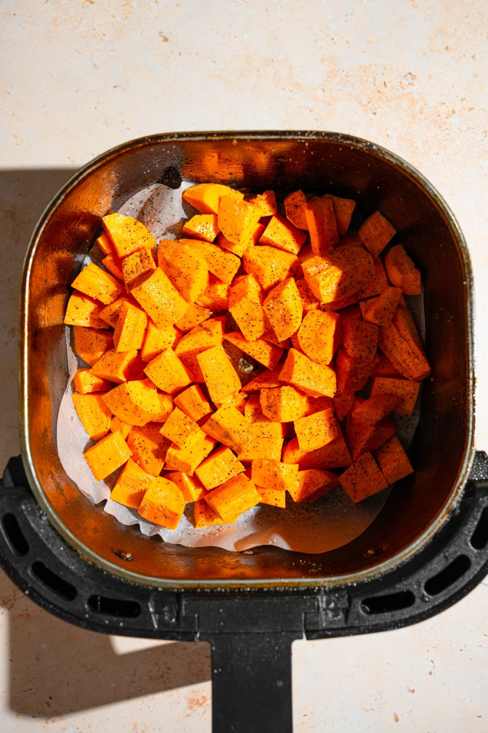 An air fryer basket lined with parchment paper with cubed sweet potatoes. The basket is on a tan counter.