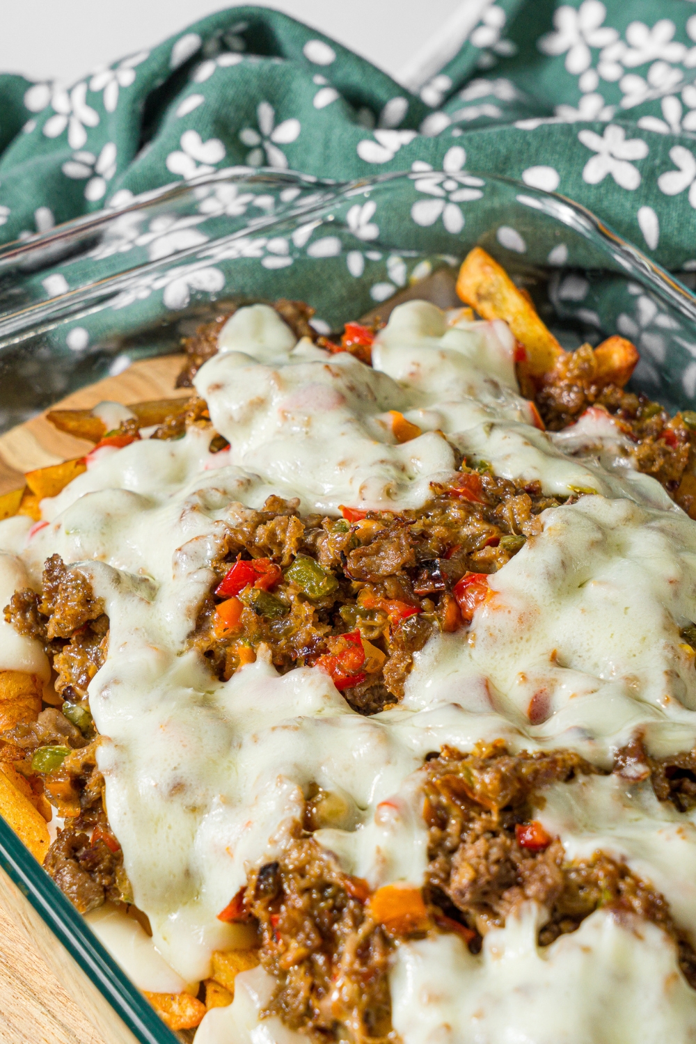 A baking dish with baked Philly cheesesteak loaded fries including fries, melted cheese, and a philly cheesesteak mixture. The dish is on a marble counter with a green floral napkin.