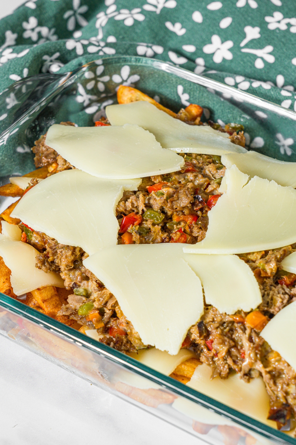 A baking dish with philly cheesesteak loaded fries mixture topped with sliced provolone cheese. The dish is on a marble counter with a green floral napkin.