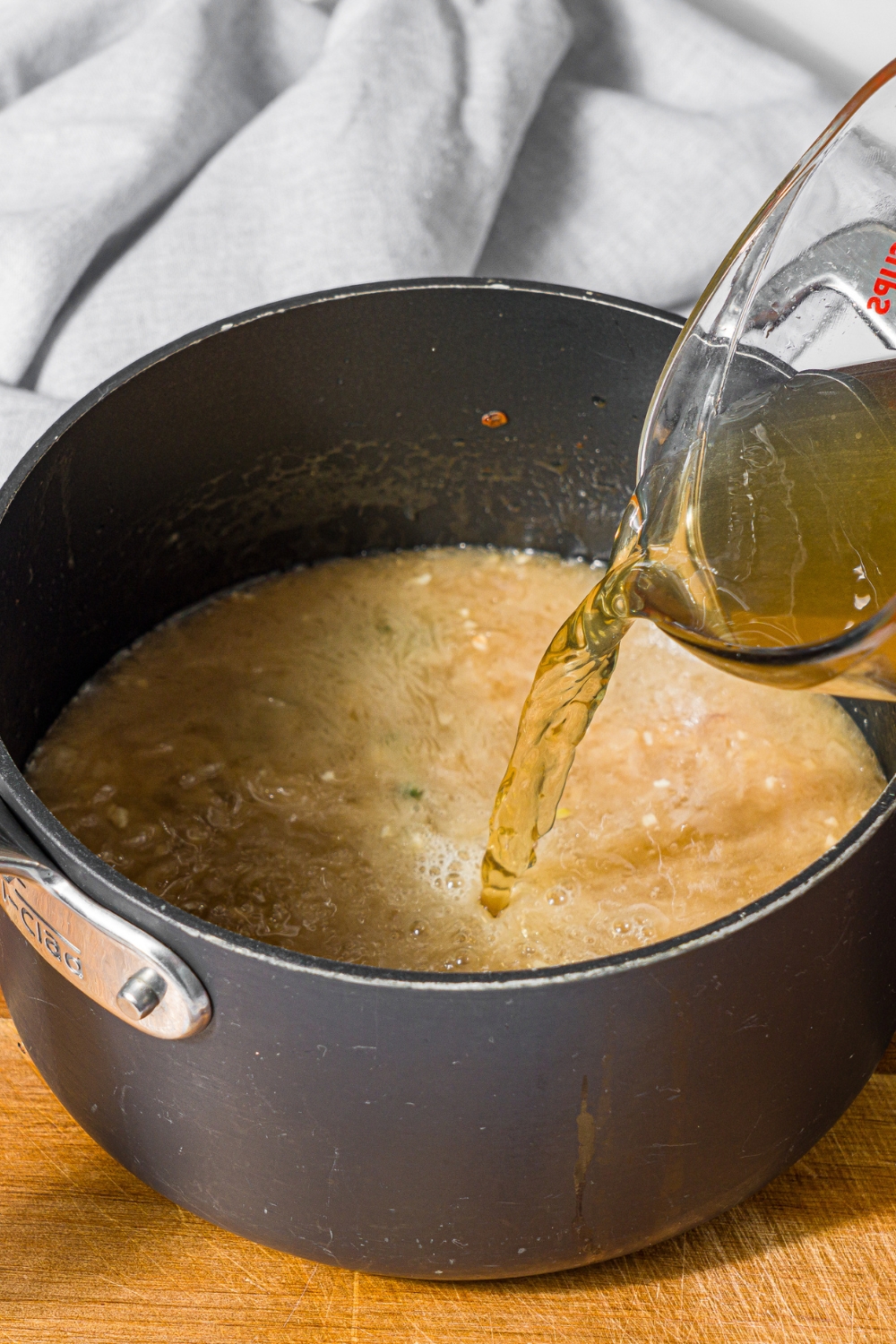 A saucepan with chicken stock being added to a steak frites sauce mixture. The saucepan is on a wooden board.