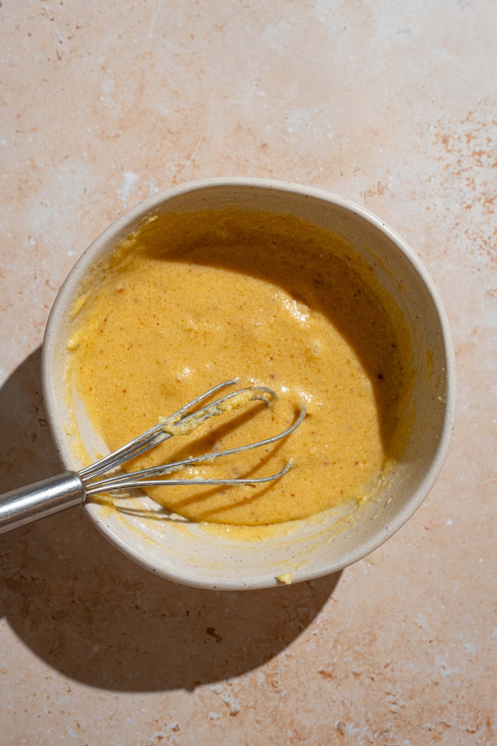 A bowl with a whisk mixing a creamed butter, egg, and vanilla mixture. The bowl is on a tan counter.
