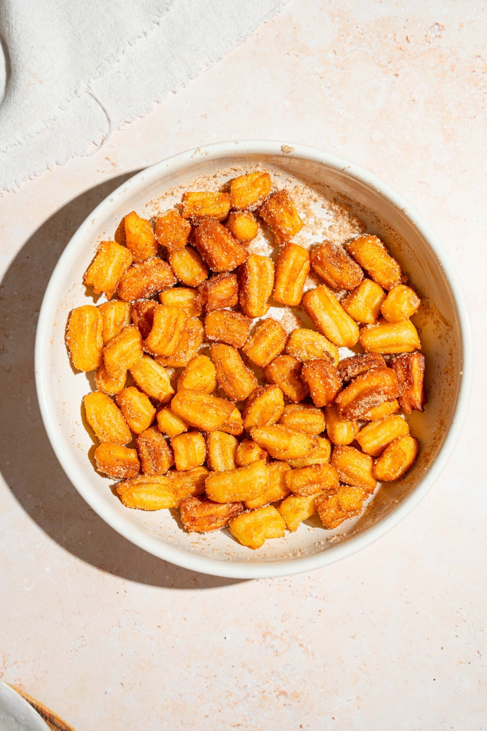 A white bowl with mini churros tossed in sugar and cinnamon. The bowl is on a tan counter.