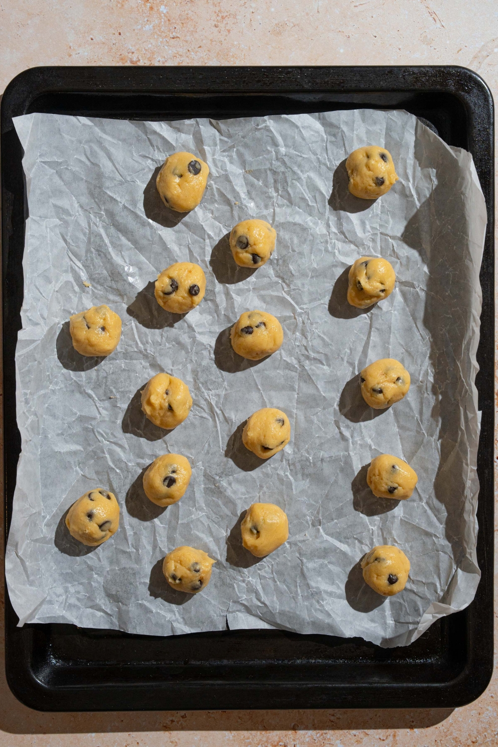 A baking sheet lined with parchment paper with mini chocolate chip cookie dough balls. The sheet is on a tan counter.