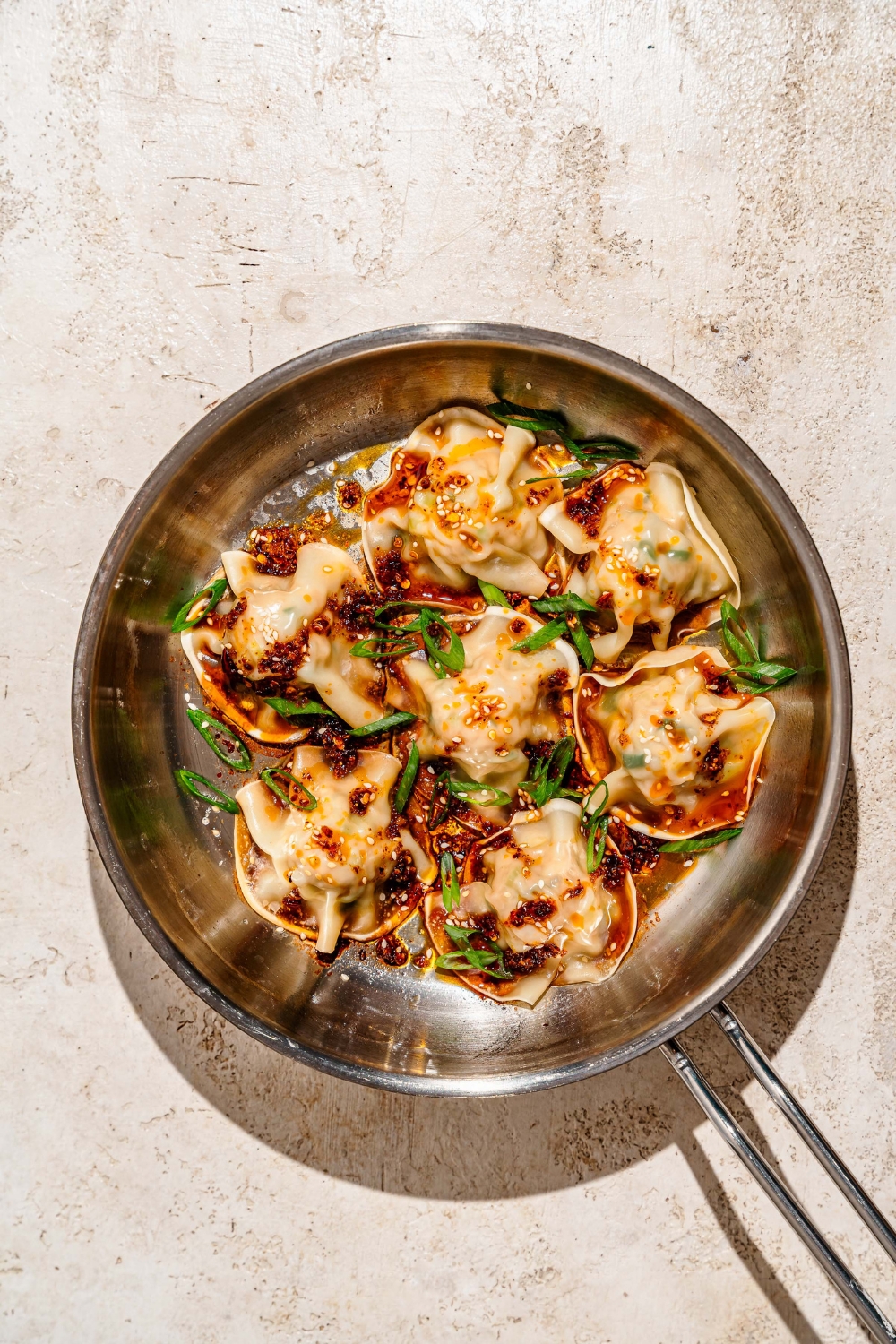 A skillet with pan fried pork and shrimp dumplings topped with sliced green onion, sesame seeds, and a soy sauce chili oil mixture. The skillet is on a tan counter.