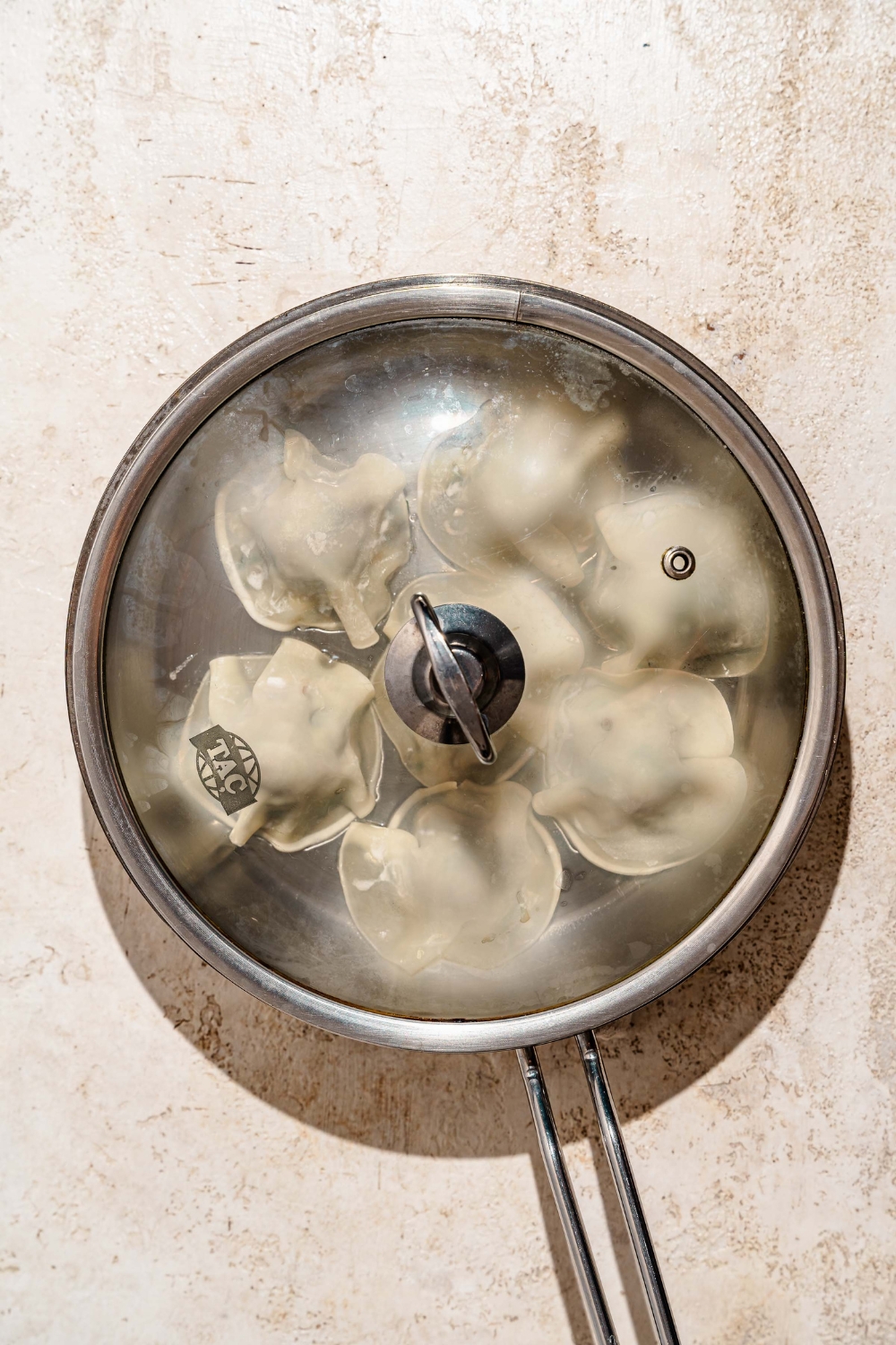A covered skillet steaming pork and shrimp dumplings. The skillet is on a tan counter.
