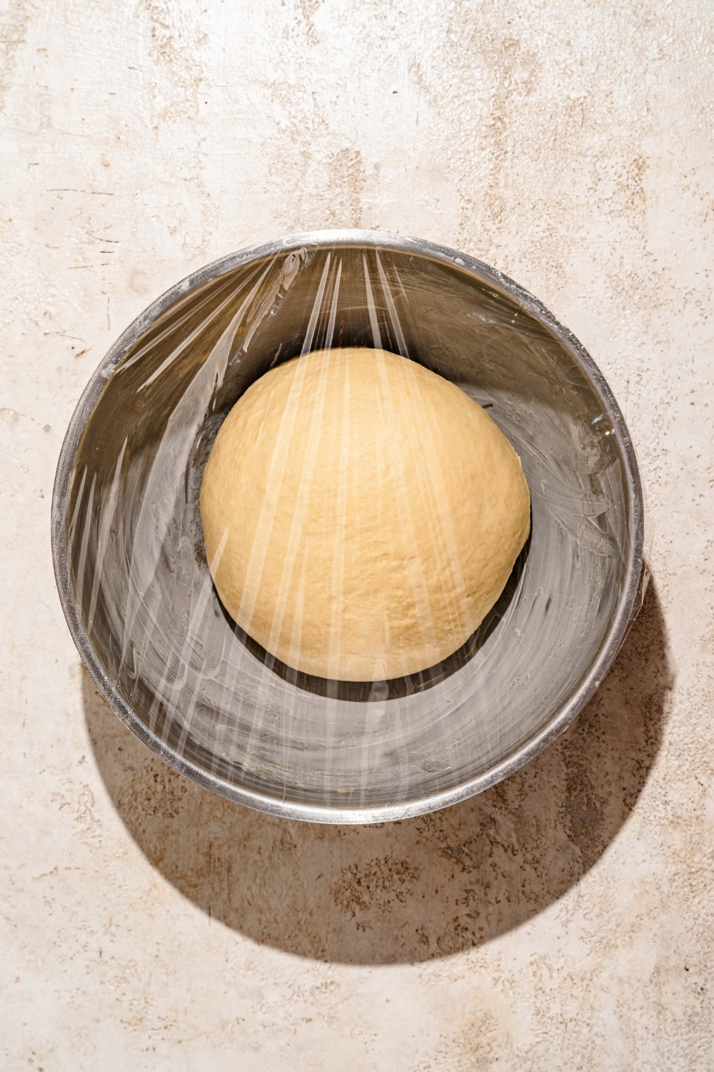 A large covered mixing bowl with a ball of sticky bun dough. The bowl is on a tan counter.