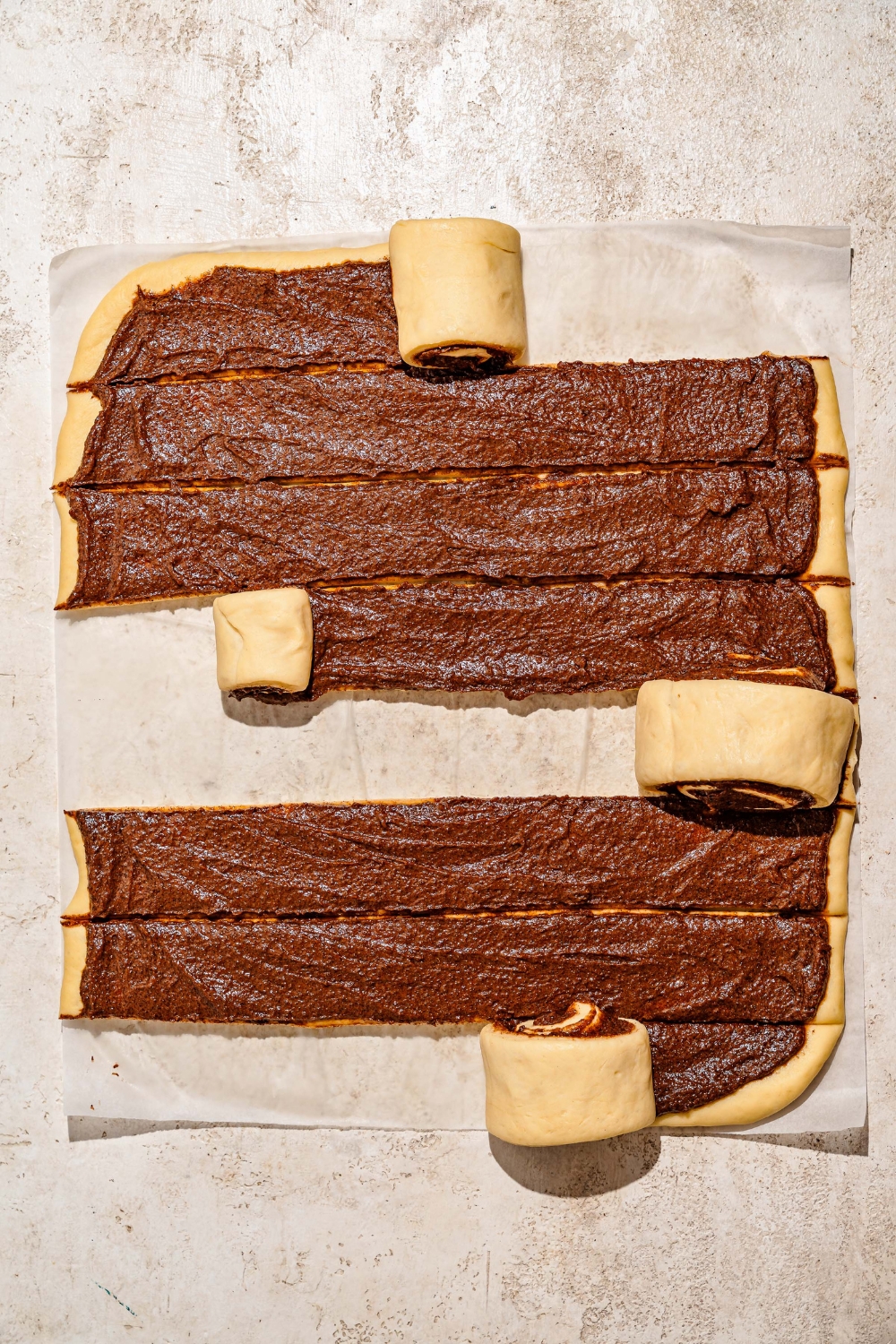A tan counter with a rolled out rectangle of sticky bun dough topped with caramel mixture and sliced in several rows. Each row is being rolled into buns.