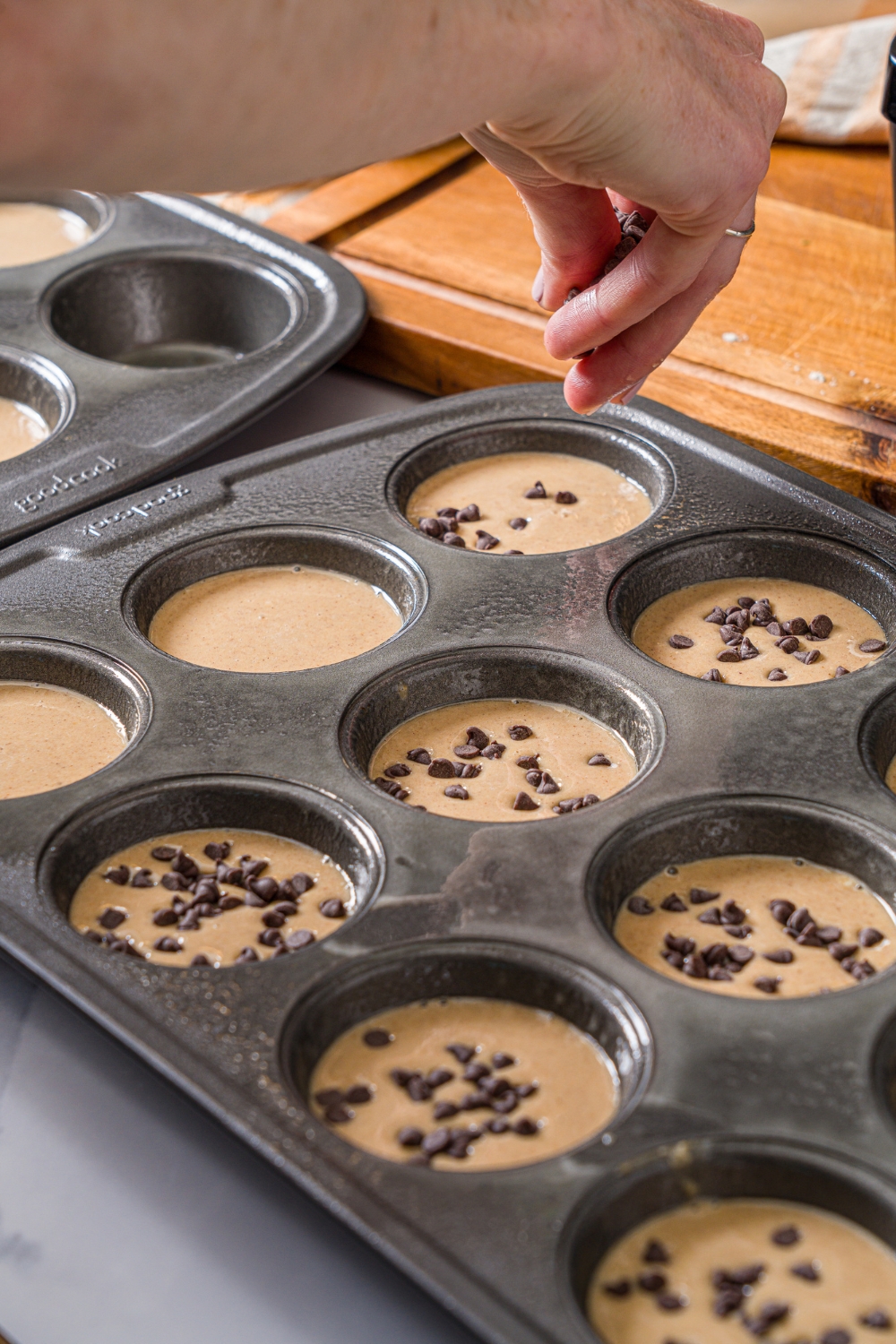 A muffin tin filled with protein banana muffin batter. A hand is adding chocolate chips to the tin. The tin is on a marble counter.
