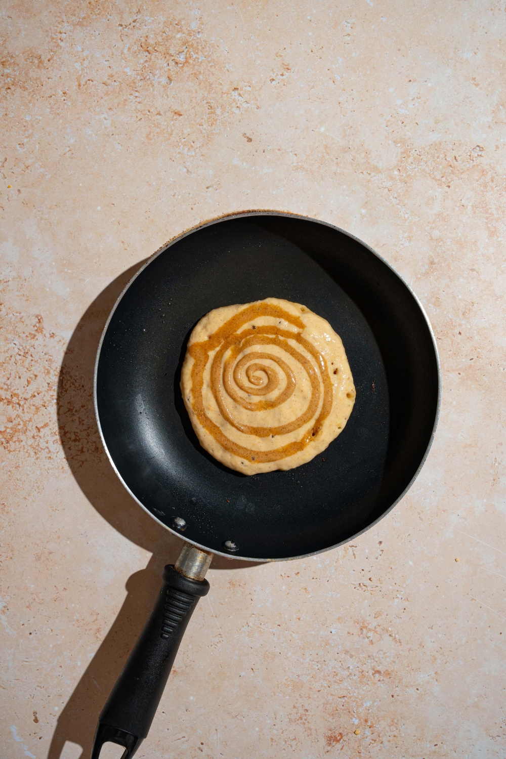 A skillet with an unflipped pumpkin pancake with cinnamon swirl. The skillet is on a tan counter.