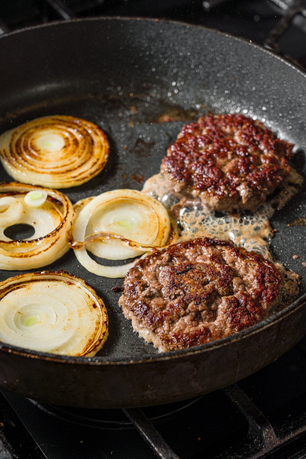 A skillet with two smashed burger patties cooking with sliced onion.