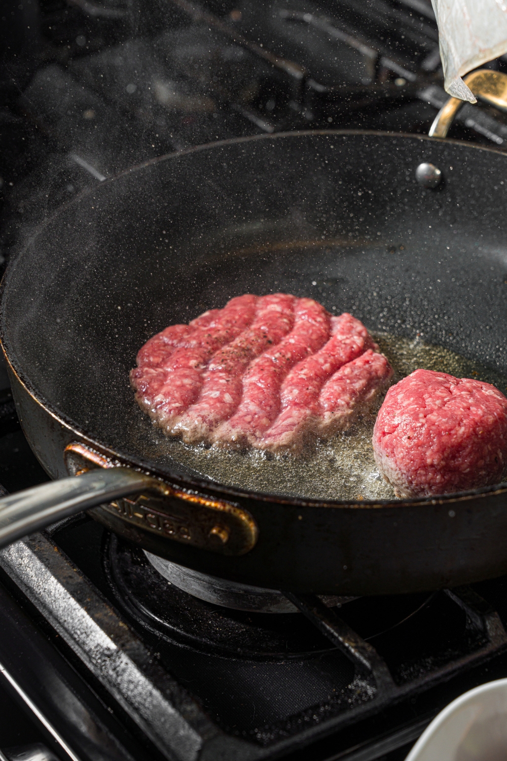 A skillet with two seasoned burger patty balls. One of the patties has been smashed by a burger press. The skillet is on a stove.