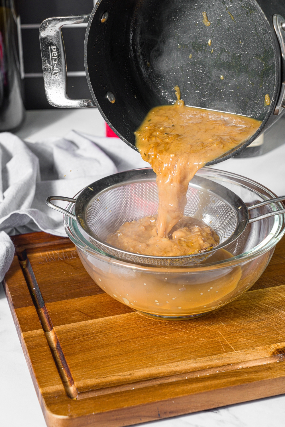 A saucepan of steak frites sauce being poured through s strainer over a glass bowl. The bowl is on a wooden board on a marble counter.