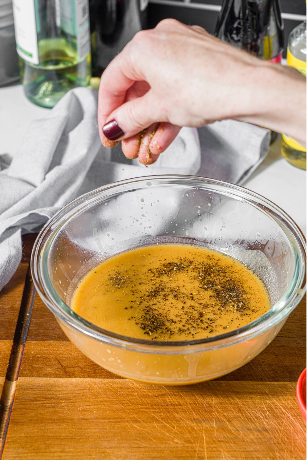 A glass bowl with seasonings being added to a strained steak frites sauce. The bowl is on a wooden board.