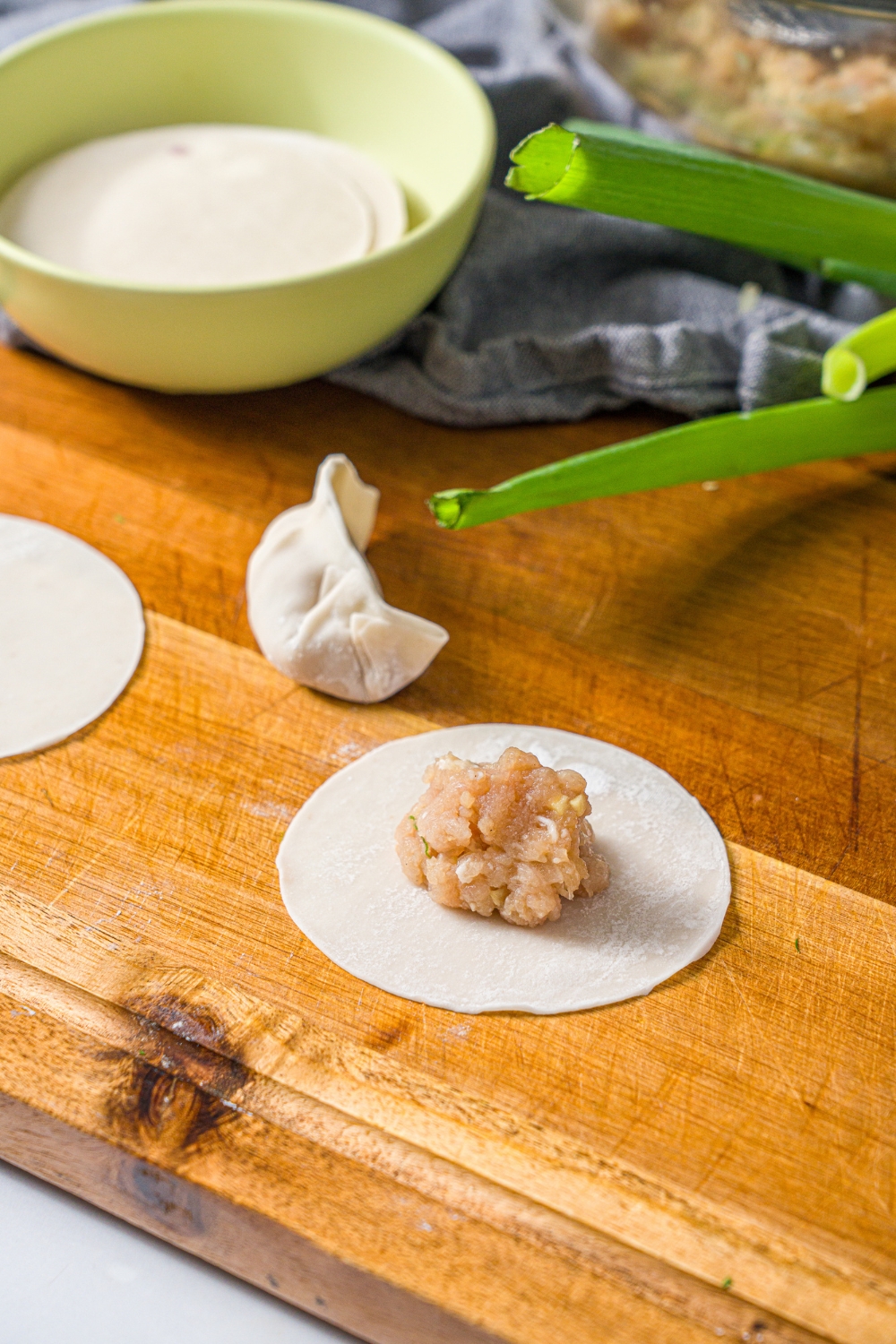 A wooden board with an open dumpling wrapper topped with a pork mixture. There is another wrapped pork dumpling on the board.
