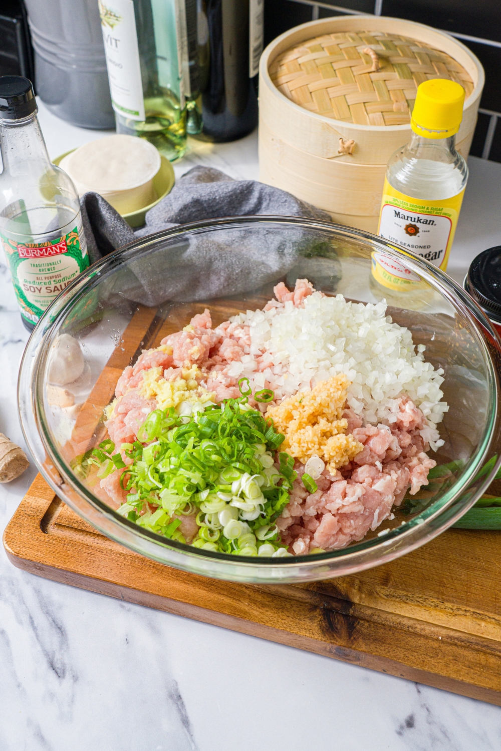 A glass bowl with ingredients to make pork mixture including ground pork, onion, garlic, ginger, and green onion. The bowl is on a wooden board.