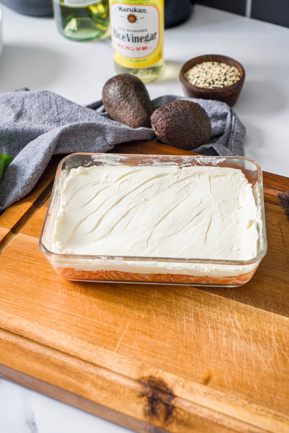 A glass baking dish layered with cubed sushi salmon topped with cream cheese. The dish is on a wooden board.