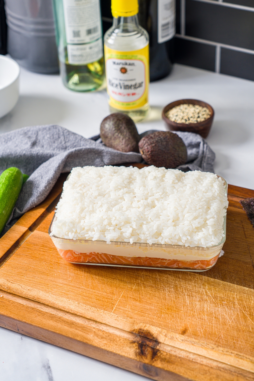 A glass baking dish layered with cubed sushi salmon topped with cream cheese and sushi rice. The dish is on a wooden board.