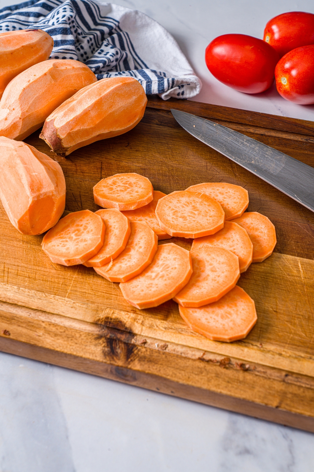 A wooden board with peeled sweet potatoes and sliced sweet potato medallions. The board is on a marble counter.