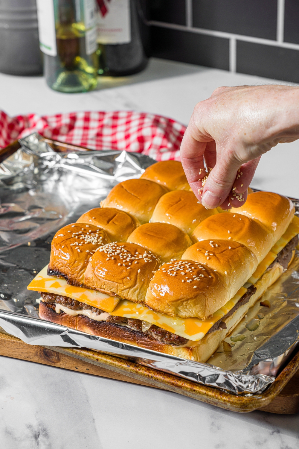 A baking sheet lined with foil with assembled cheeseburger sliders with a hand sprinkling sesame seeds on top. The sheet is on a wooden board.
