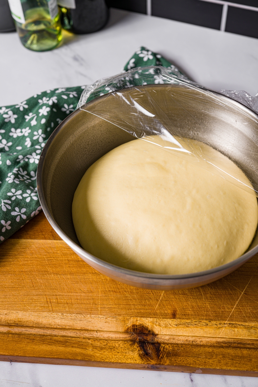 A large bowl partially covered with saran wrap with a ball of beignet dough that has been proofed. The bowl is on a wooden board.