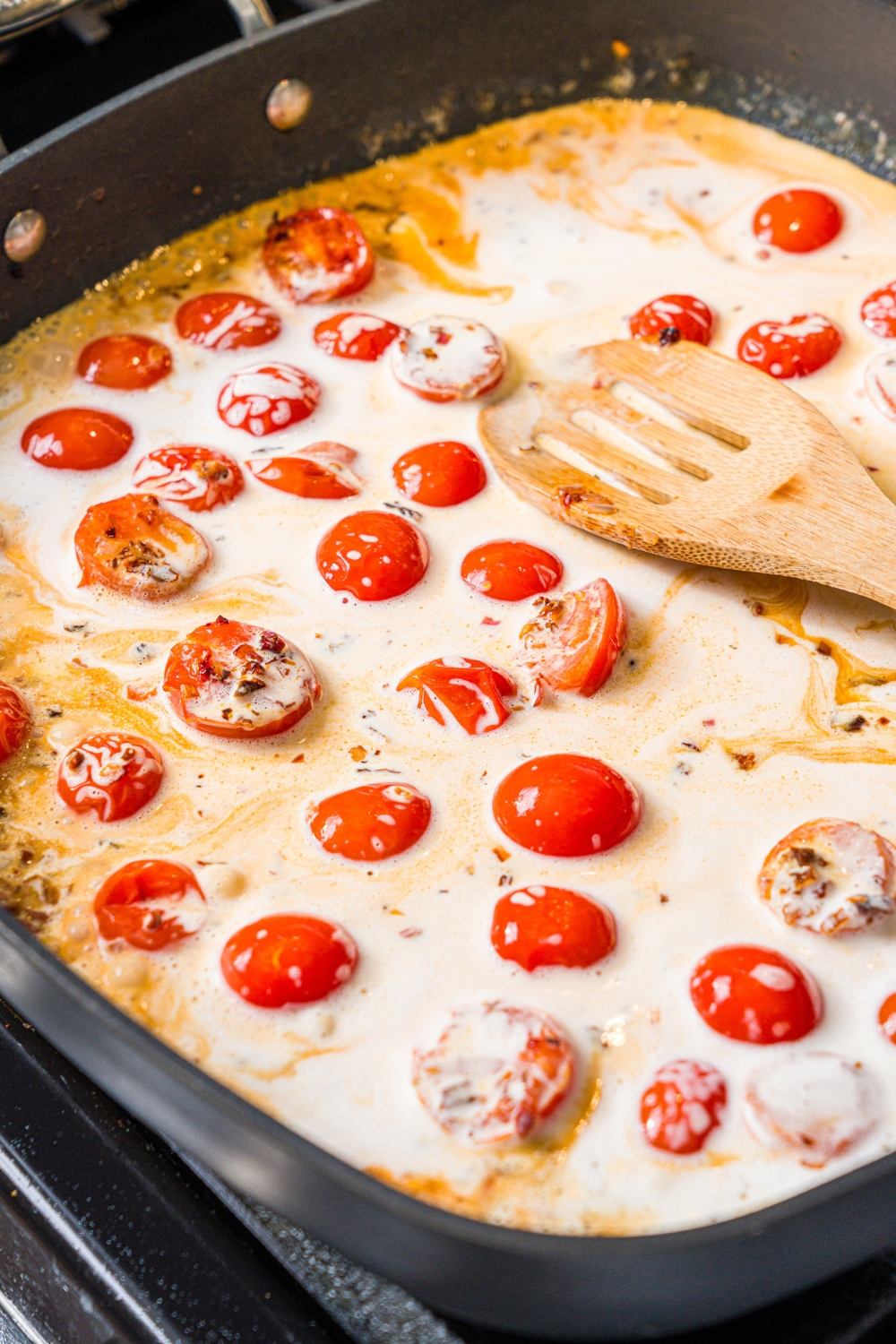 A skillet with a wooden spoon stirring a cream sauce mixture with sautéed garlic and tomatoes. The skillet is cooking on the stove.
