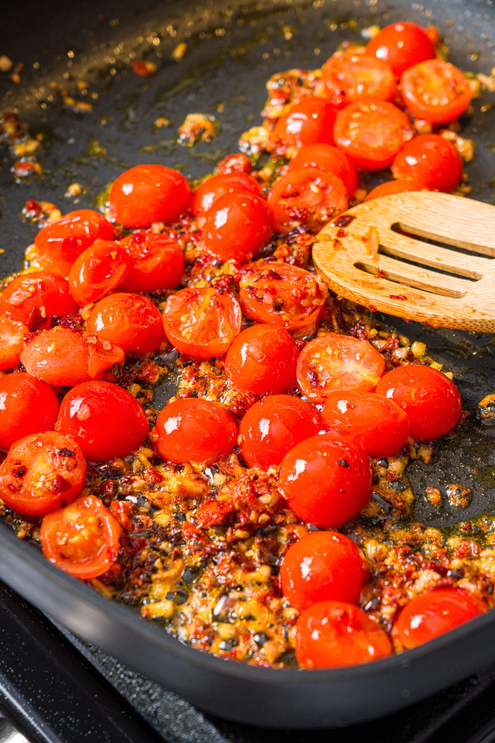 A skillet with a wooden spoon stirring a sautéed tomatoes and garlic mixture. The skillet is cooking on the stove.