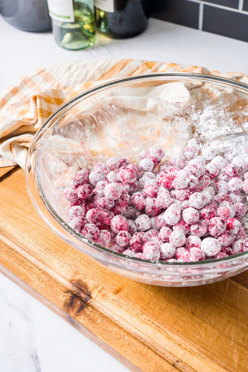 A large glass bowl with soaked cranberries tossed in sugar. The bowl is on a wooden board with a yellow striped napkin.
