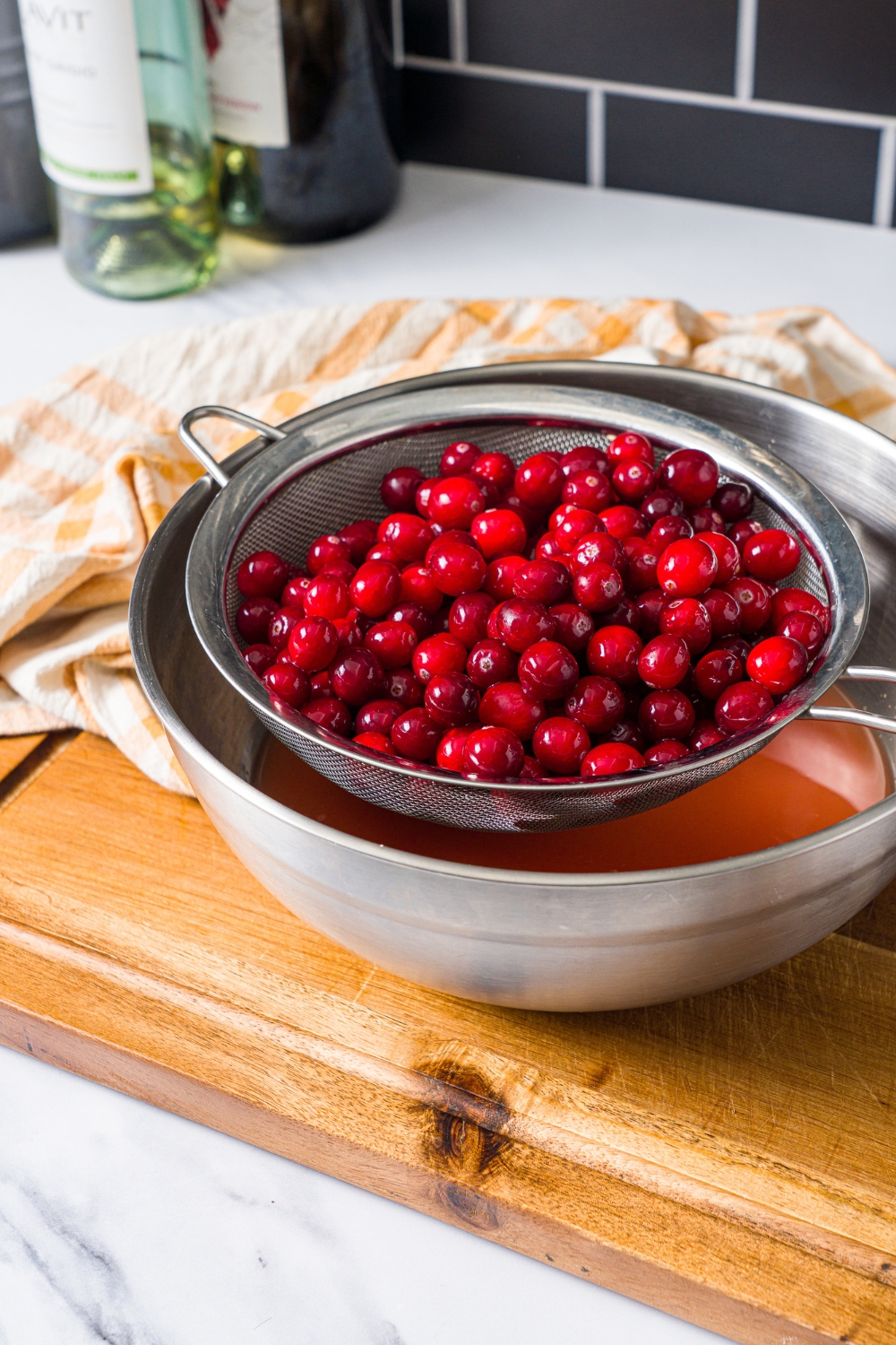 Cranberries being strained through a mesh strainer over a large bowl on a wooden board.