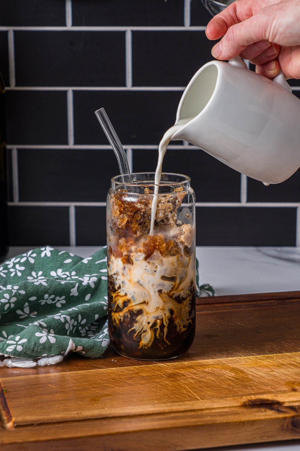 A glass of coffee slushie with milk being poured in the glass. The glass is on a wooden board.