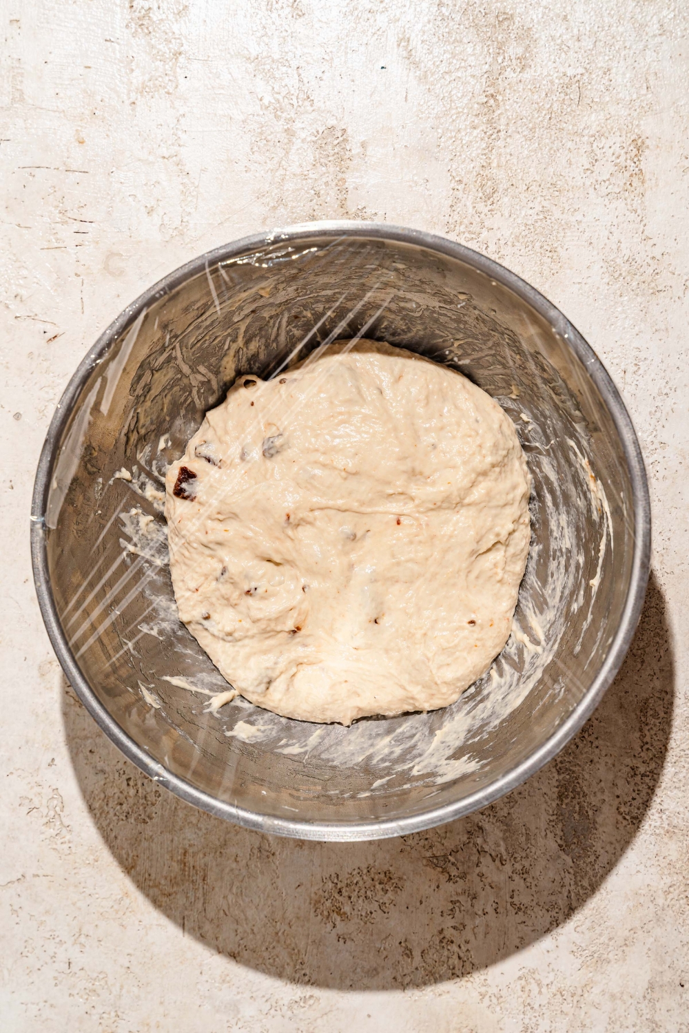 A large bowl with a focaccia dough wrapped in saran wrap. The bowl is on a tan counter.