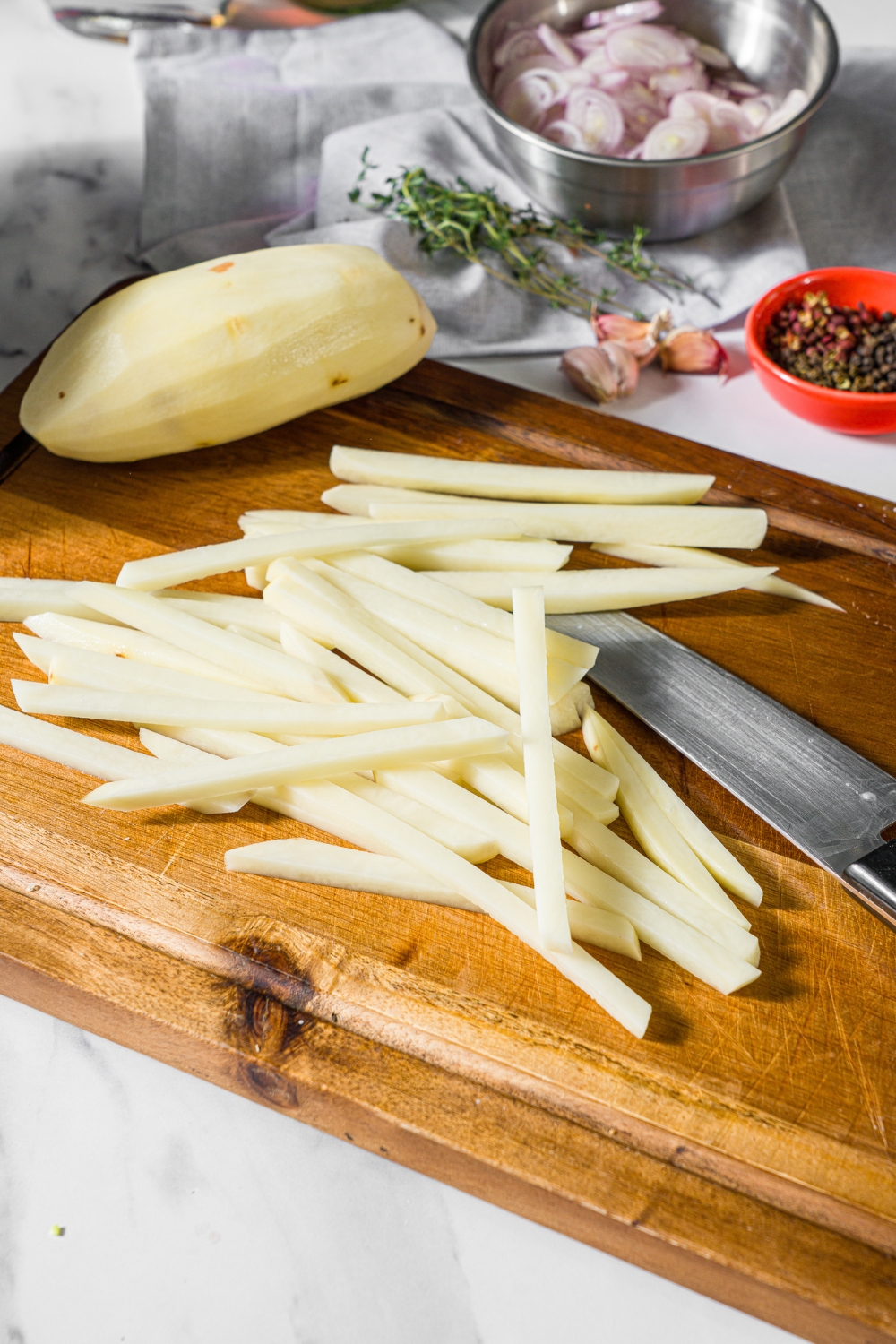 A wooden cutting board with slices of potato cut into fries. There is a knife on the board.