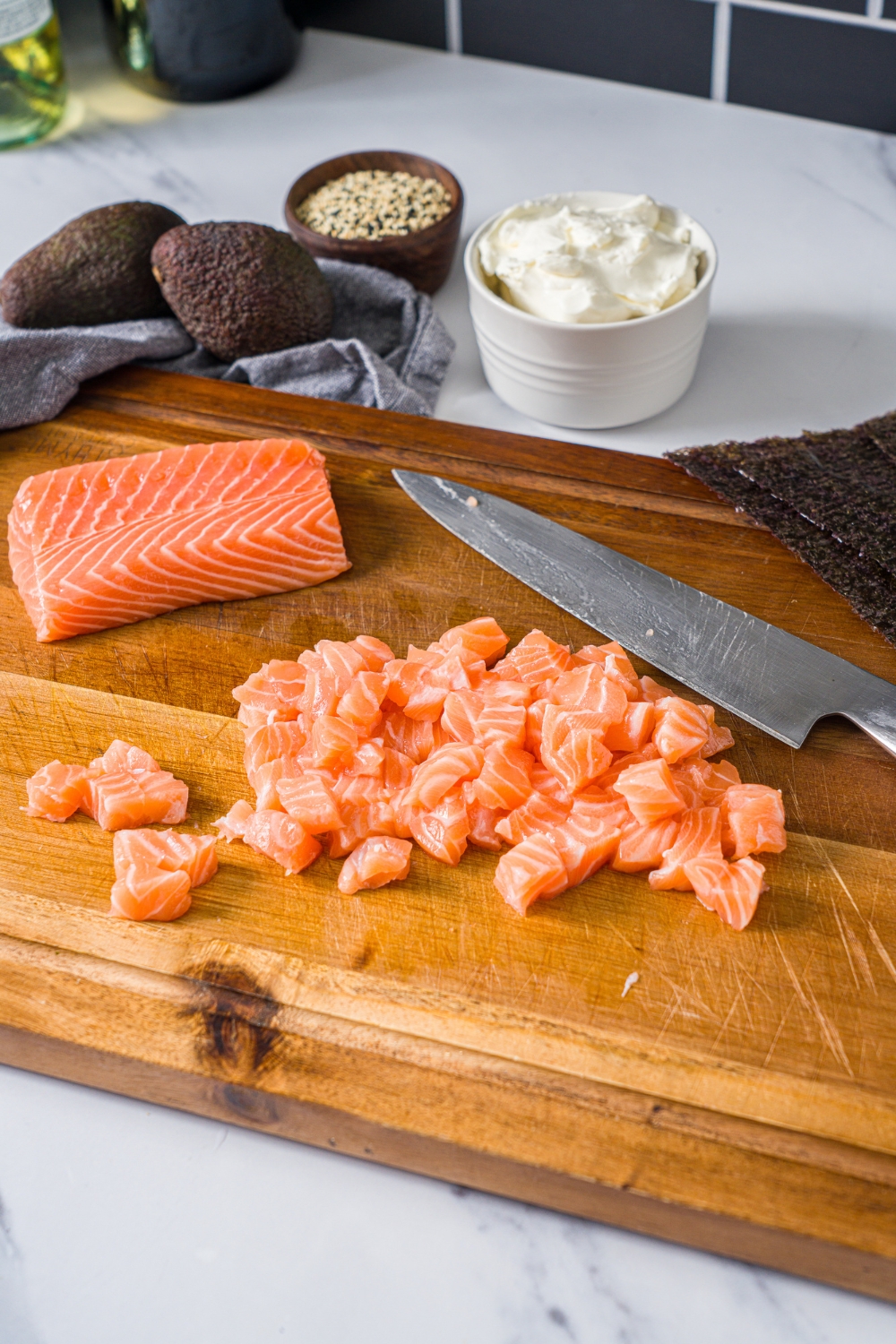 A wooden cutting board with cubed salmon and a salmon filet. There is a knife on the board. The board is on a marble counter with avocado and a bowl of cream cheese.