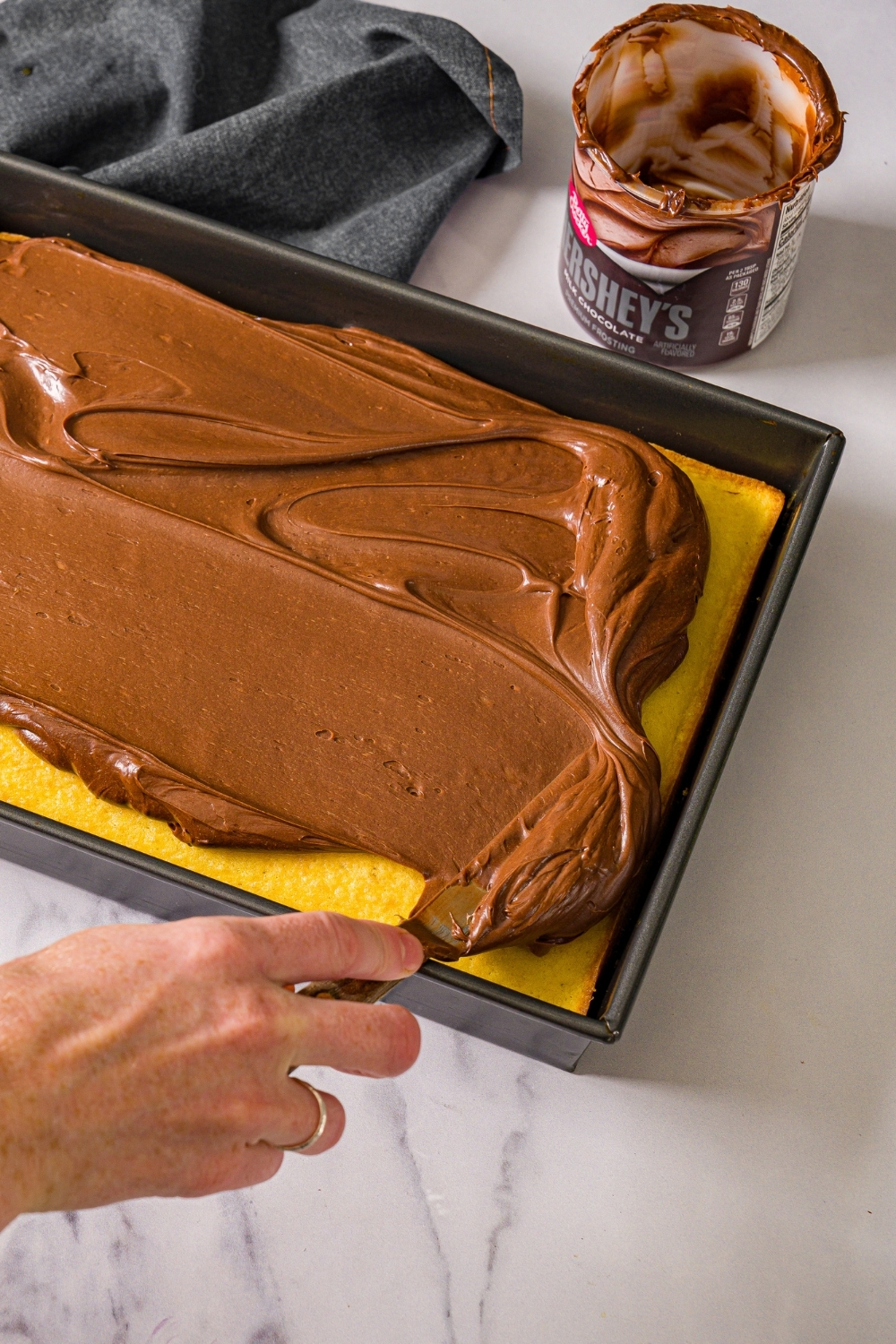 A baking pan with yellow cake and a frosting knife spreading chocolate frosting over the cake. The pan is on a marble counter with a container of frosting.