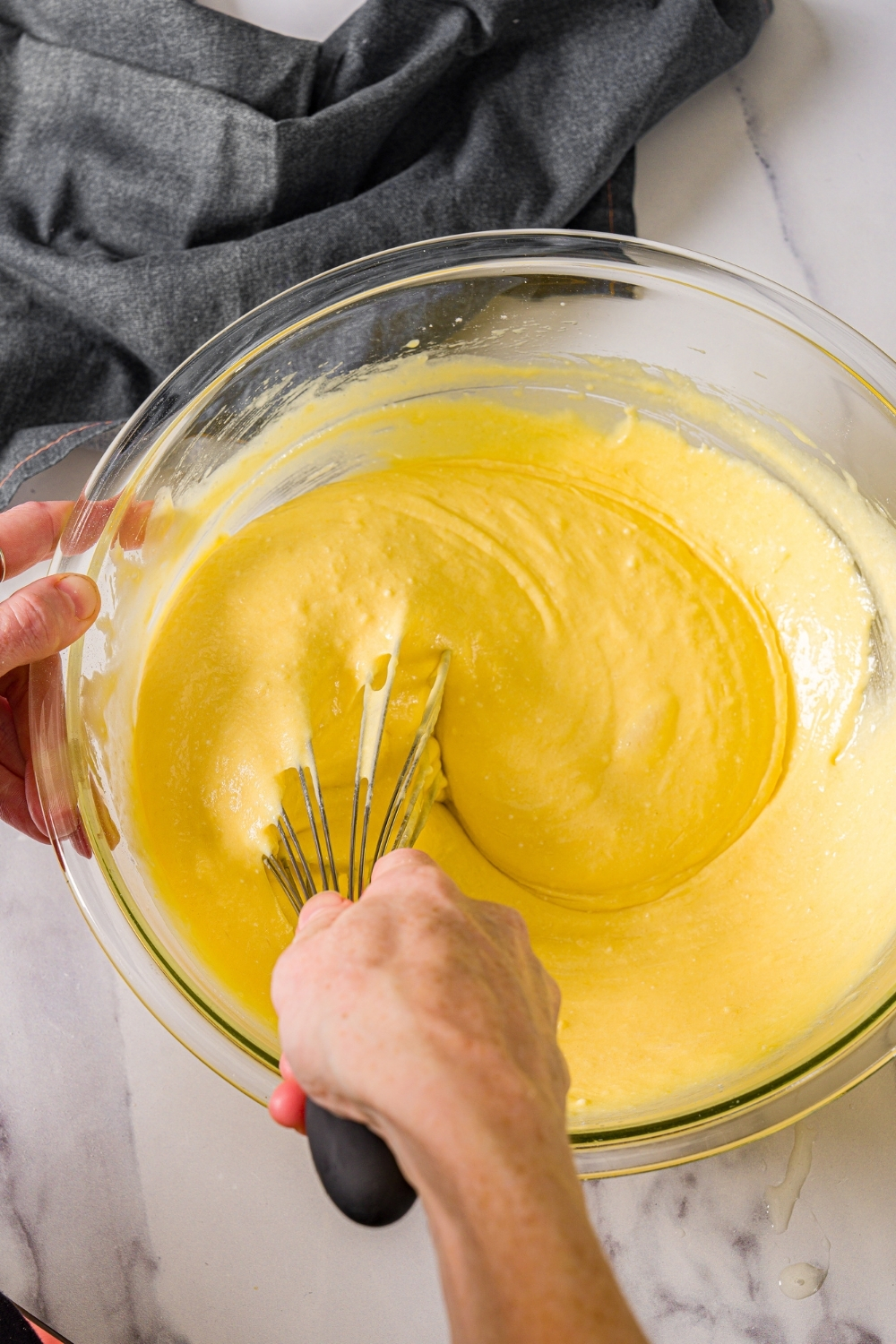 A large mixing bowl with a whisk mixing yellow cake batter. The bowl is on a marble counter with a gray cloth napkin.